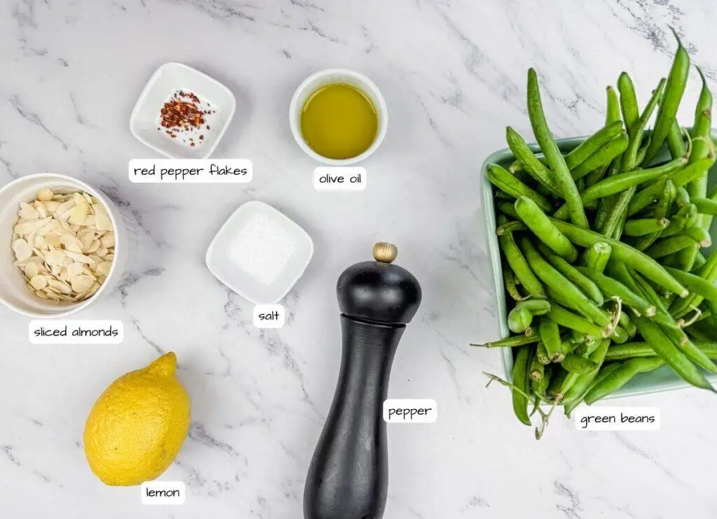 Ingredients on a marble surface: sliced almonds, red pepper flakes, olive oil, salt, pepper grinder, lemon, and green beans in a green container.
