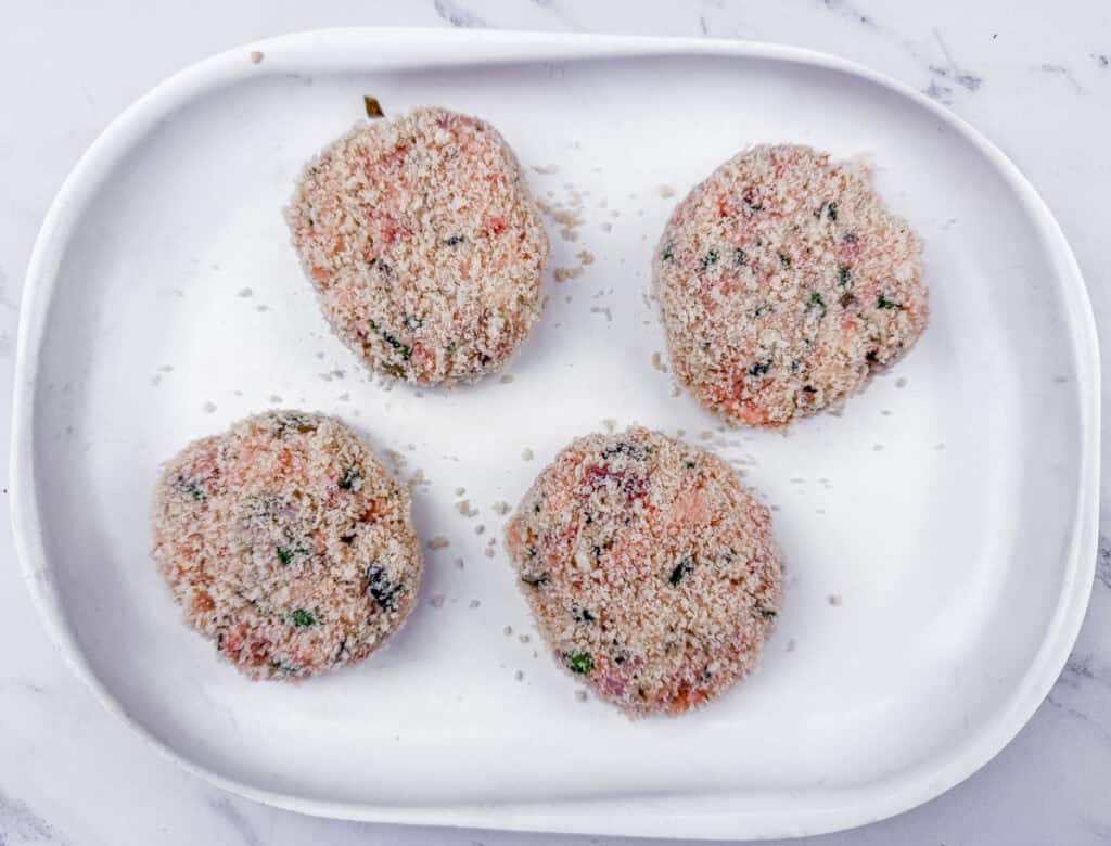 Four uncooked, breaded patties on a white oval plate, placed on a marble surface.