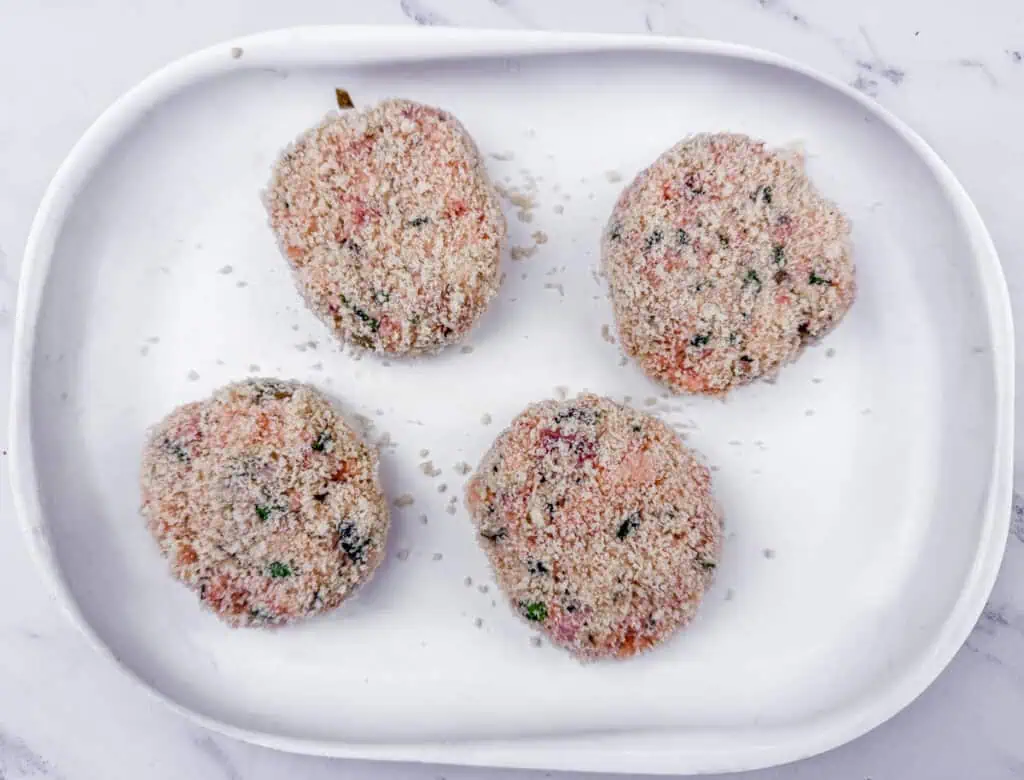 Four uncooked, breaded patties on a white oval plate, placed on a marble surface.