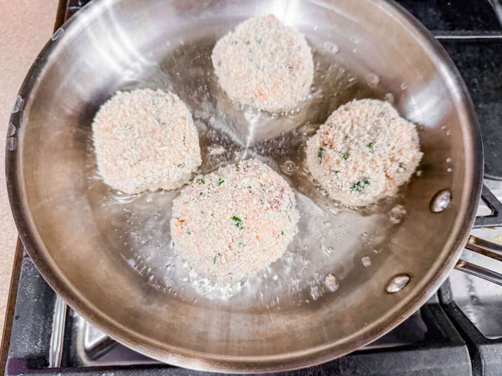 Four breaded patties frying in a stainless steel pan on a stove.