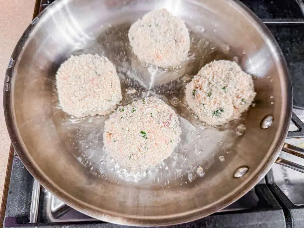 Four breaded patties frying in a stainless steel pan on a stove.