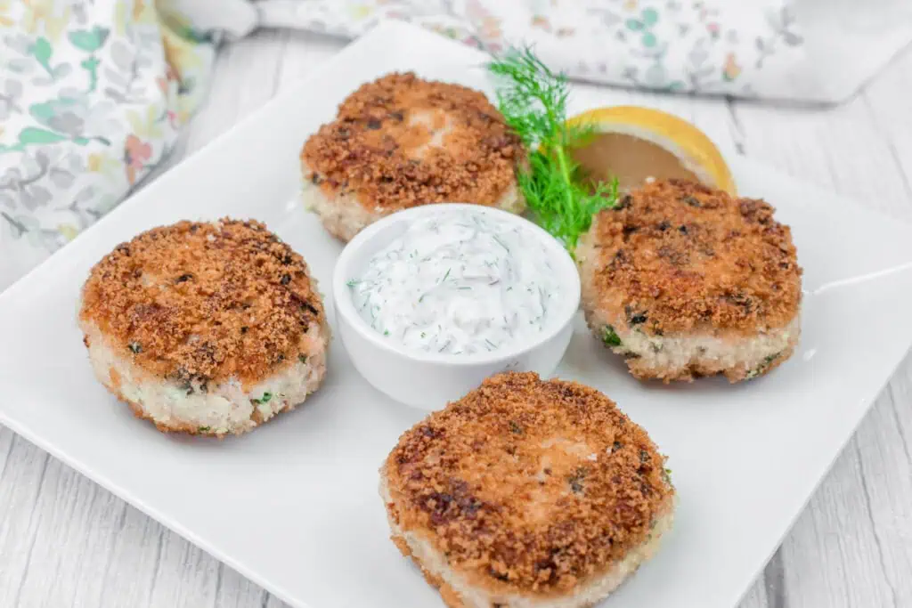 Four Salmon Cakes on a square white plate with a small dish of creamy sauce. Lemon slice and dill garnish are present.