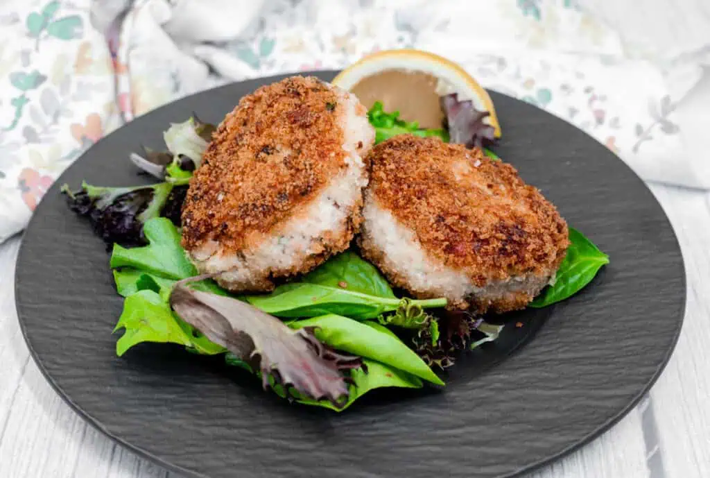 Two Salmon Cakes on a bed of mixed greens with a lemon wedge on a black plate.