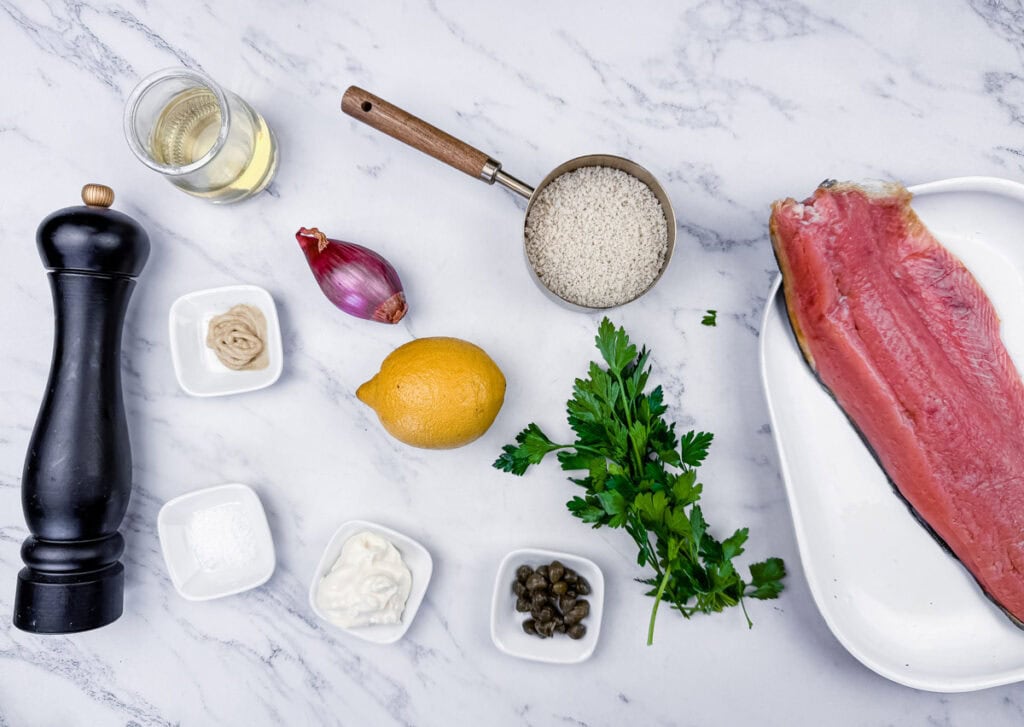 Ingredients arranged on a marble surface: uncooked fish fillet, rice, wine, lemon, shallot, parsley, pepper grinder, mustard, mayonnaise, and capers.