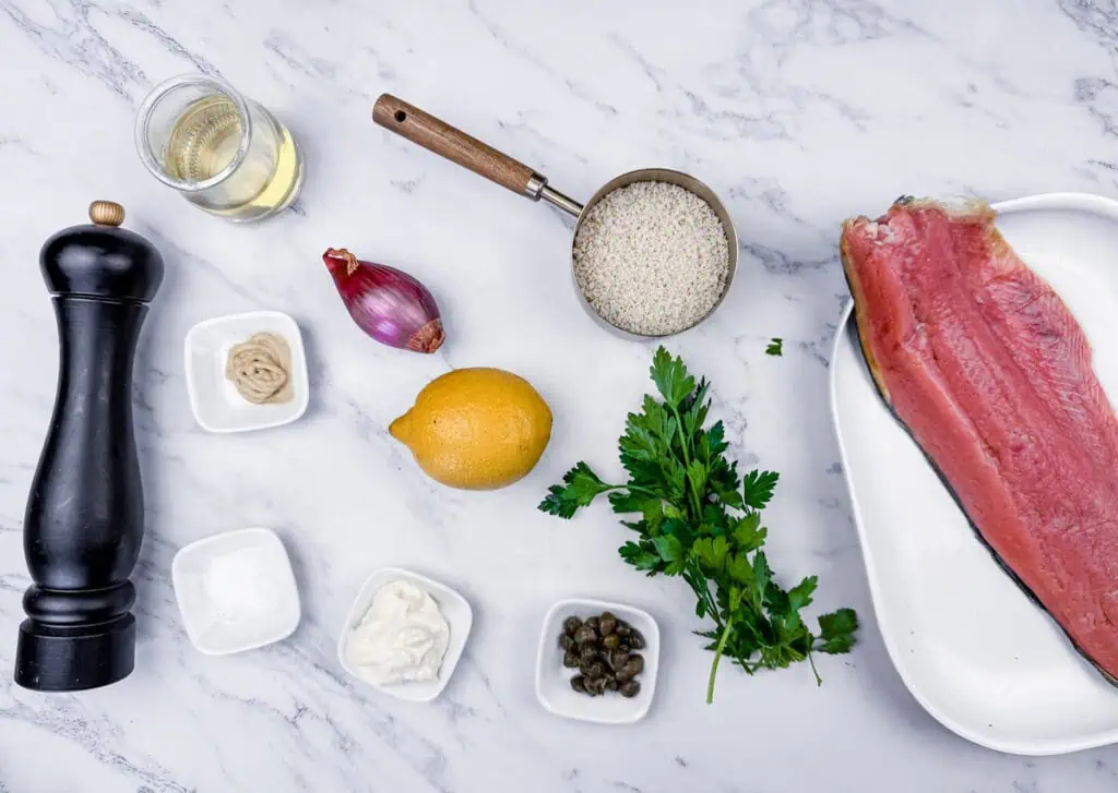 Ingredients arranged on a marble surface: uncooked fish fillet, rice, wine, lemon, shallot, parsley, pepper grinder, mustard, mayonnaise, and capers.