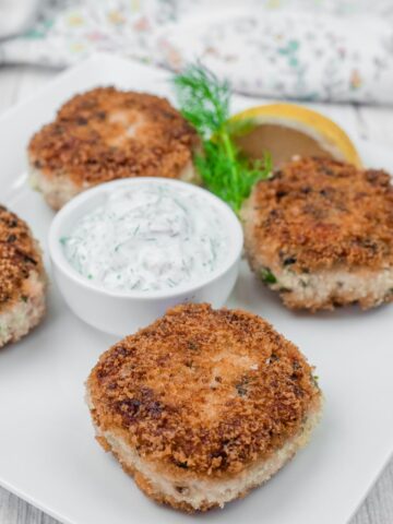 Four Salmon Cakes with a side of creamy dill sauce and a lemon wedge on a white plate.