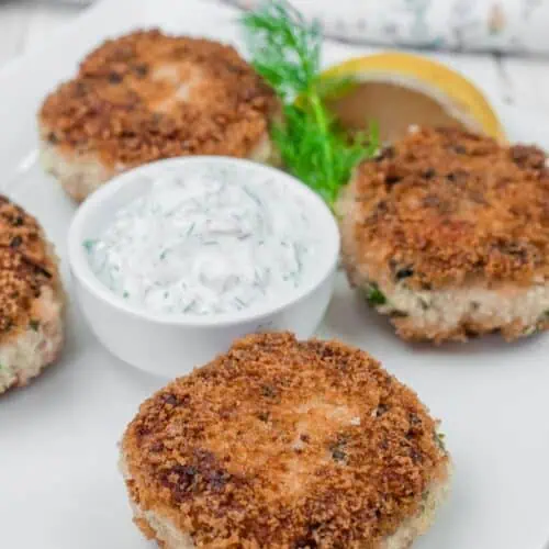 Four Salmon Cakes with a side of creamy dill sauce and a lemon wedge on a white plate.