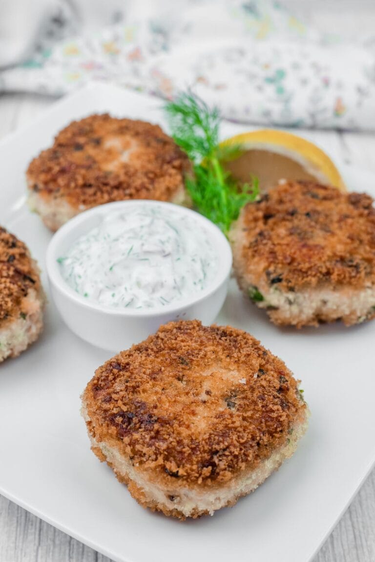 Four Salmon Cakes with a side of creamy dill sauce and a lemon wedge on a white plate.