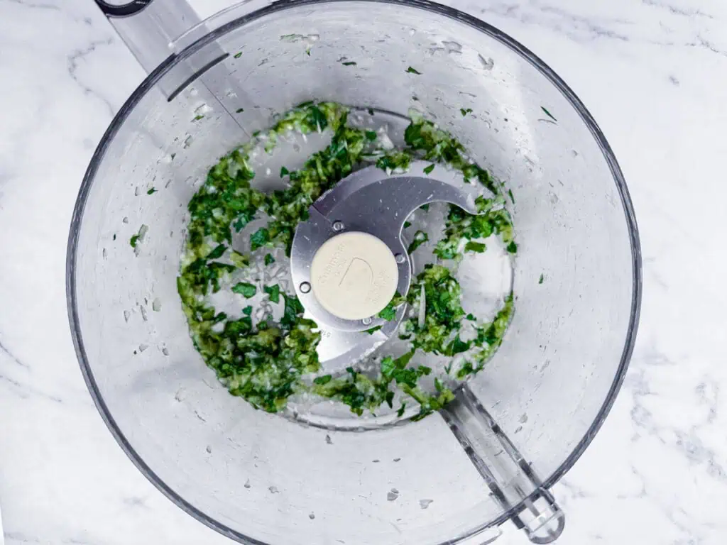 Overhead view of a food processor bowl with chopped green herbs and garlic on a marble countertop.