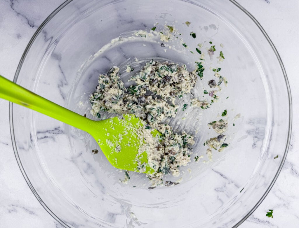 A green spatula in a clear bowl containing a mixture of chopped ingredients. The bowl rests on a white marble surface.