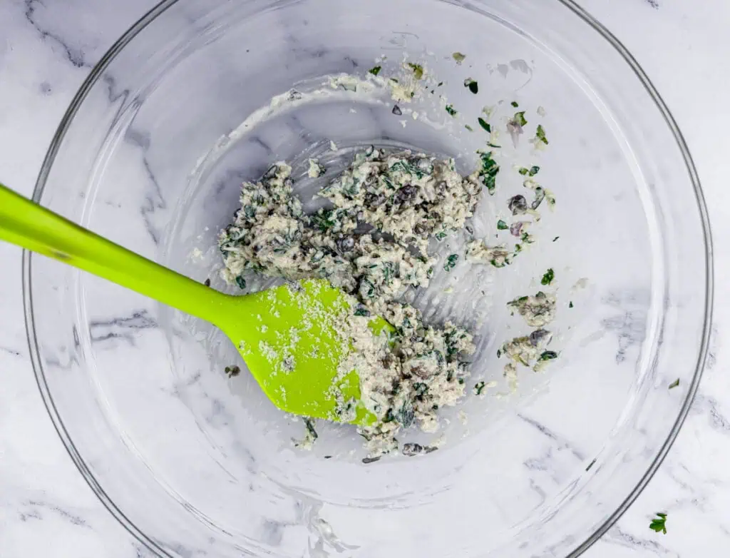 A green spatula in a clear bowl containing a mixture of chopped ingredients. The bowl rests on a white marble surface.