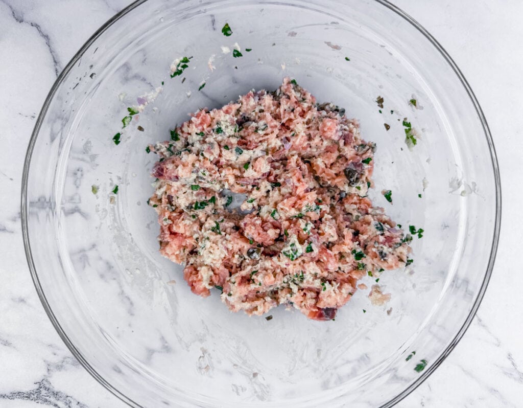 A glass bowl containing a mixed raw meat mixture with herbs.