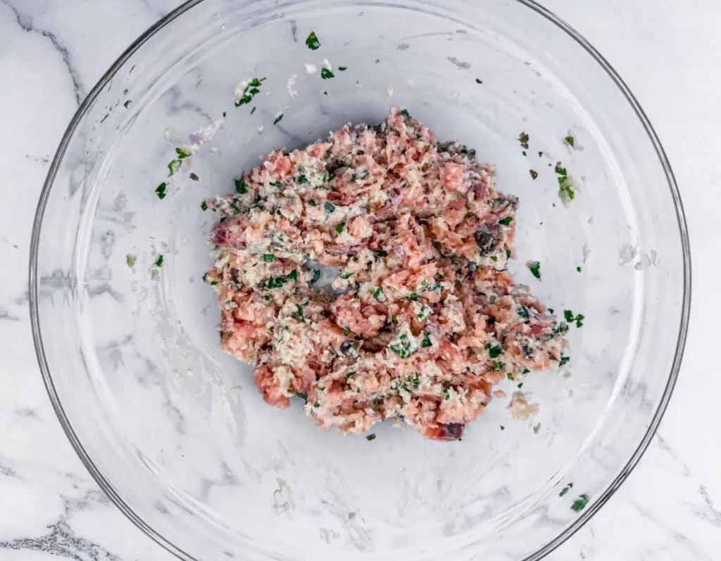 A glass bowl containing a mixed raw meat mixture with herbs.