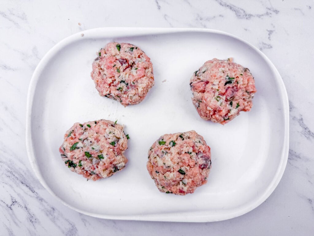 Four uncooked meat patties with herbs on a white oval plate, placed on a white marble surface.