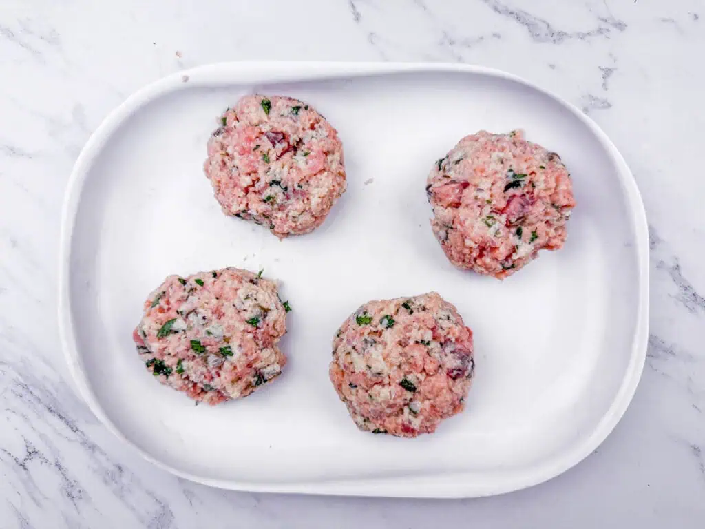 Four uncooked meat patties with herbs on a white oval plate, placed on a white marble surface.