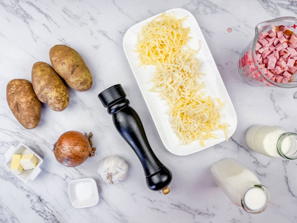 Ingredients on a marble counter: potatoes, shredded cheese on a plate, diced ham in a measuring cup, onion, garlic, butter, salt, pepper grinder, and milk in a bottle.