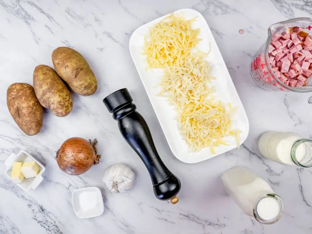 Ingredients on a marble counter: potatoes, shredded cheese on a plate, diced ham in a measuring cup, onion, garlic, butter, salt, pepper grinder, and milk in a bottle.