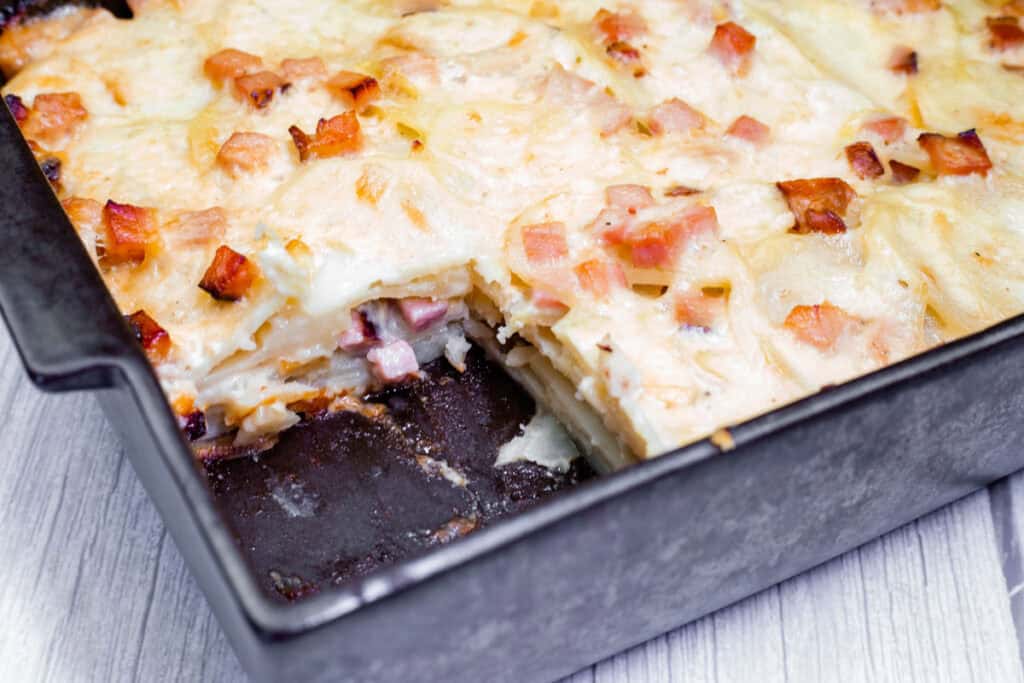 A rectangular metal baking dish with sliced Scalloped Potatoes with Ham and Cheese. A portion is missing, revealing layers of potatoes and bits of ham. The dish is on a wood-textured surface.