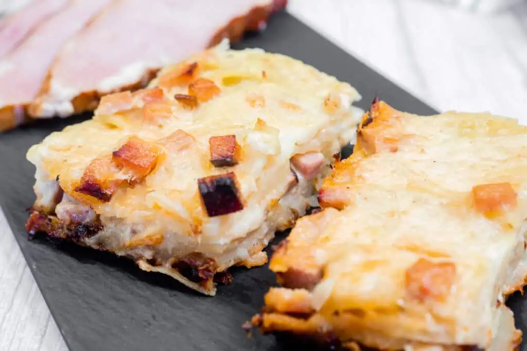 Close-up of two slices of Scalloped Potatoes with Ham and Cheese, topped with browned cheese and cubed ham, with slices of ham in the background.