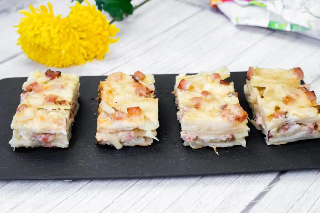 Four slices of Scalloped Potatoes with Ham and Cheese on a black slate board, with yellow flowers and a colorful napkin in the background.
