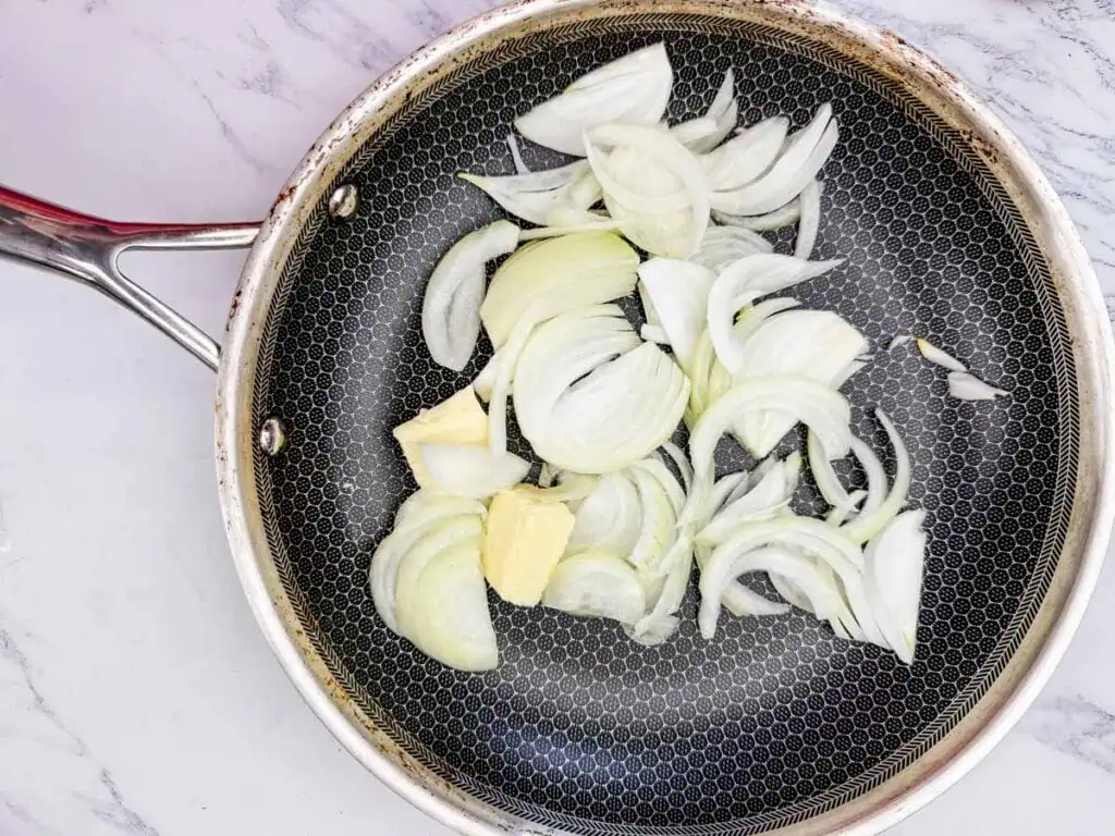 Sliced onions and butter in a frying pan on a marble countertop.