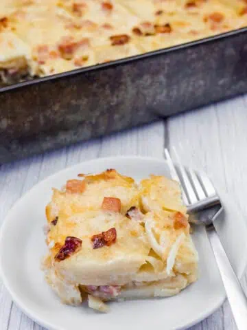 A serving of creamy Scalloped Potatoes with Ham and Cheese, topped with melted cheese and crispy browned bits, served on a white plate with a fork. The casserole dish is visible in the background on a rustic wooden surface.