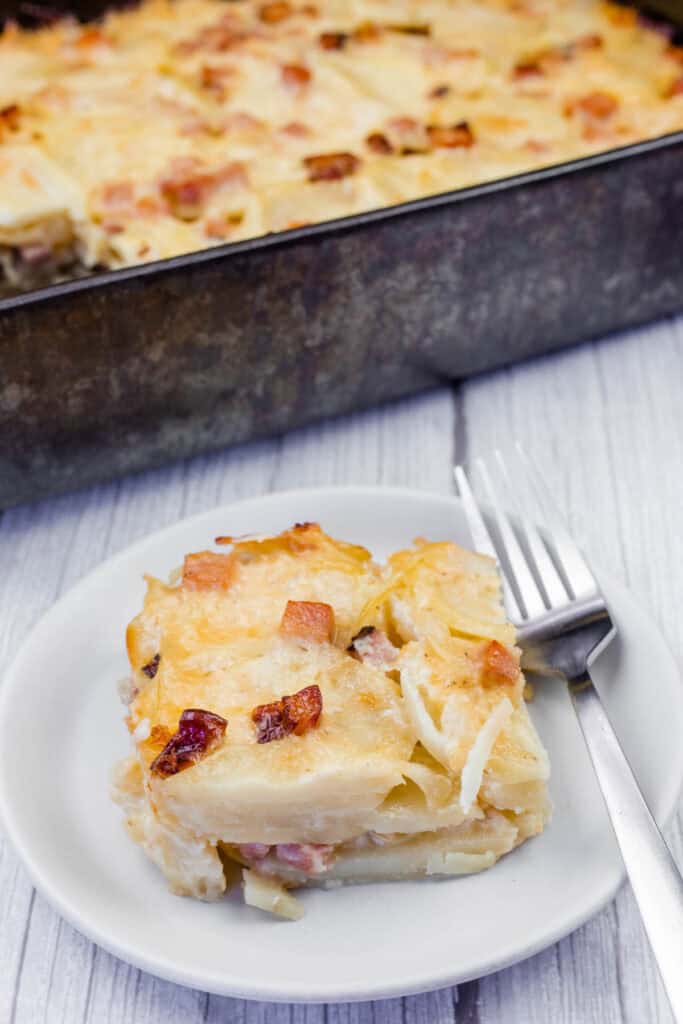 A serving of creamy Scalloped Potatoes with Ham and Cheese, topped with melted cheese and crispy browned bits, served on a white plate with a fork. The casserole dish is visible in the background on a rustic wooden surface.