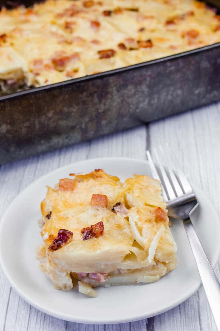 A serving of creamy Scalloped Potatoes with Ham and Cheese, topped with melted cheese and crispy browned bits, served on a white plate with a fork. The casserole dish is visible in the background on a rustic wooden surface.