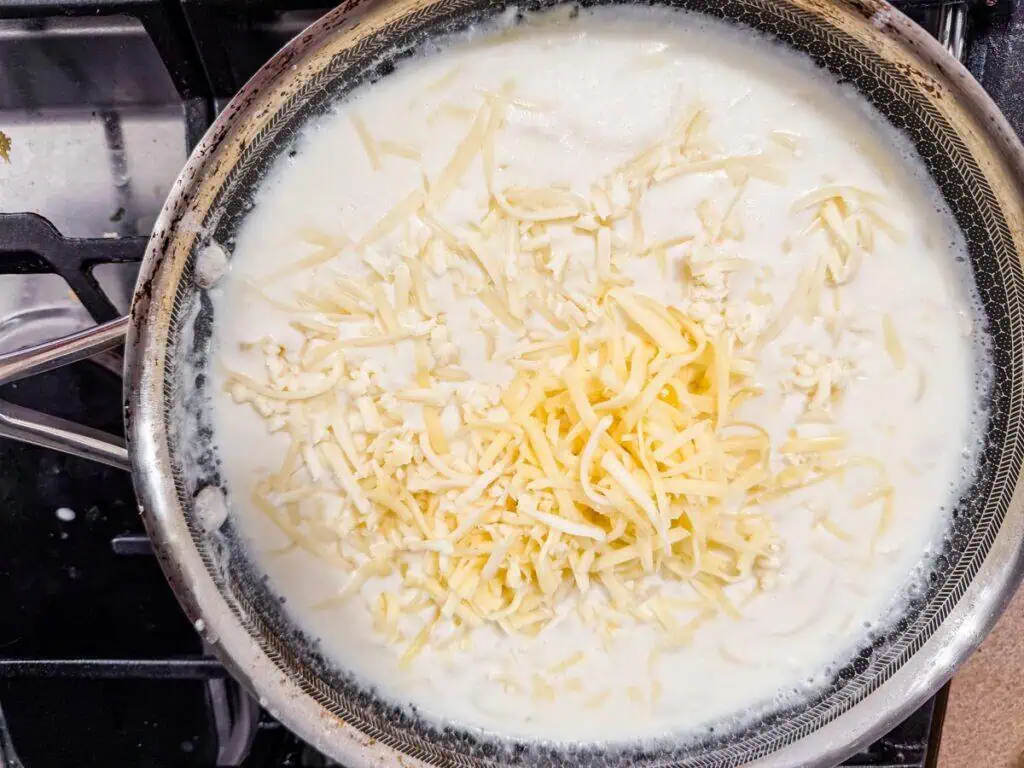 A frying pan on a stove containing a white liquid with shredded cheese on top, ready for cooking.