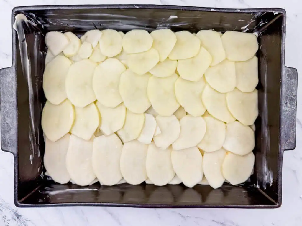 Sliced potatoes layered in a rectangular baking dish on a marble surface, ready for cooking.