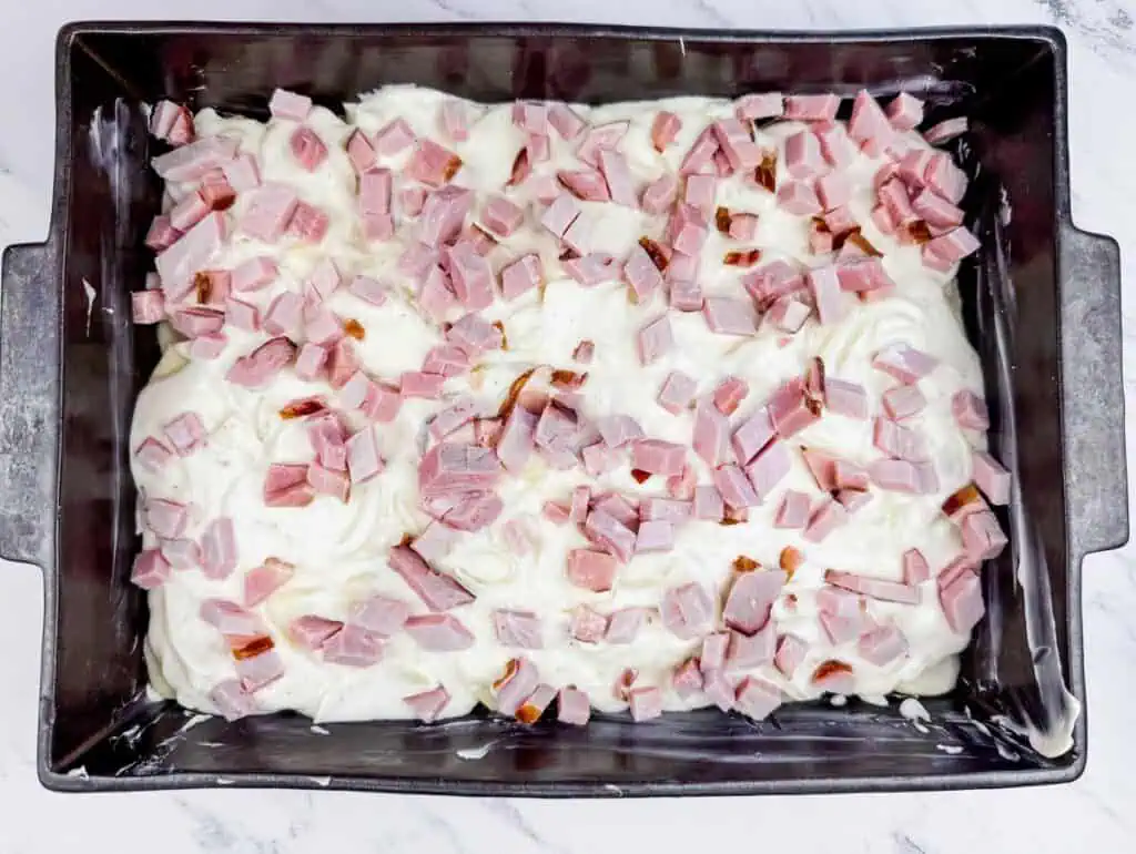 A rectangular baking dish filled with a creamy mixture topped with diced pieces of pink ham on a marble countertop.