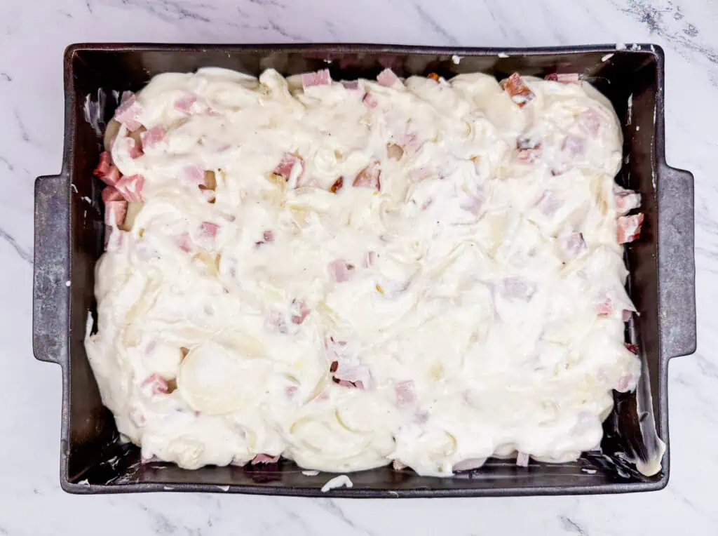 A rectangular baking dish filled with a creamy white mixture, topped with diced ham, on a marble countertop.