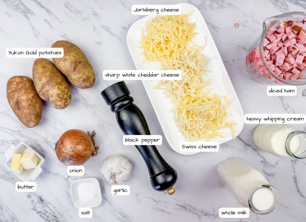 Ingredients for a dish on a marble surface: Yukon Gold potatoes, onion, garlic, butter, salt, black pepper, cheeses, diced ham, heavy cream, and whole milk.