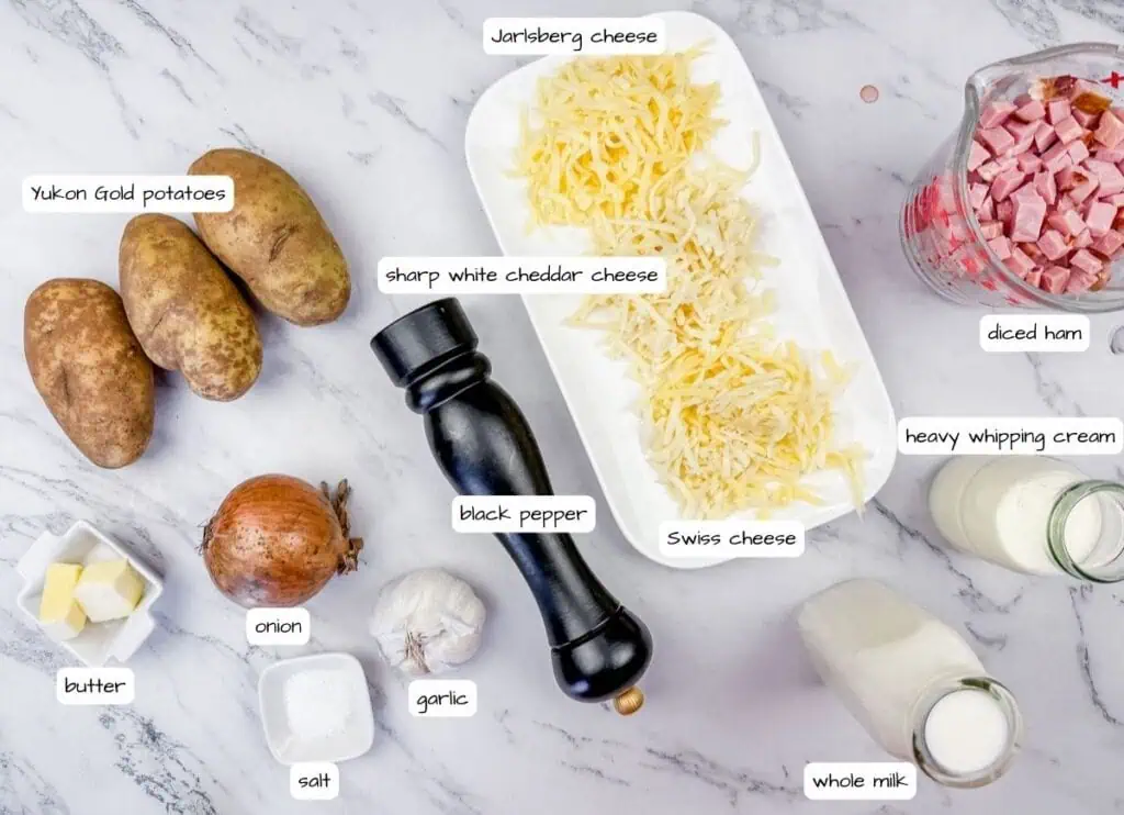 Ingredients for a dish on a marble surface: Yukon Gold potatoes, onion, garlic, butter, salt, black pepper, cheeses, diced ham, heavy cream, and whole milk.