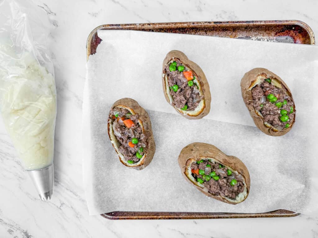 Four baked potatoes filled with ground beef, peas, and carrots on a parchment-lined tray. A piping bag with a star nozzle is nearby.