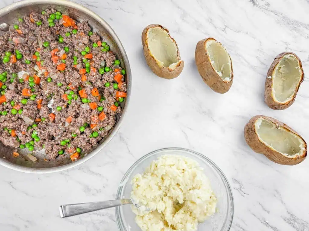 Skillet with ground beef, carrots, and peas; a bowl of mashed potatoes with a spoon; four hollowed potato skins on a marble surface.