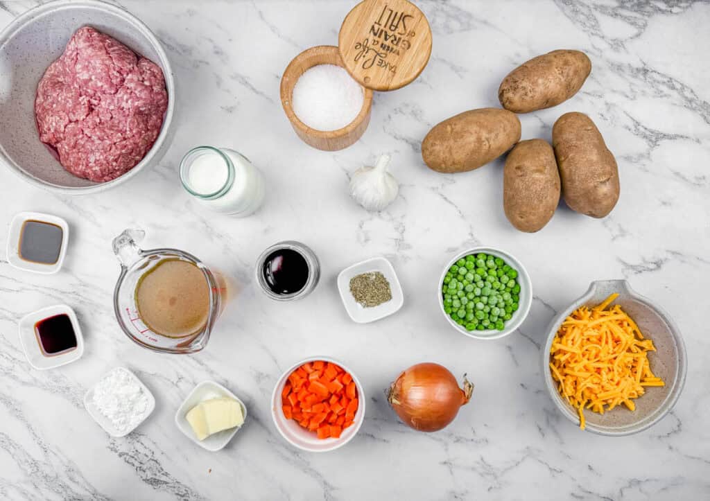 Ingredients for Shepherd&rsquo;s Pie Baked Potatoes arranged on a marble surface, including ground meat, potatoes, onion, carrots, peas, butter, broth, cream, soy sauce, garlic, salt, pepper, cornstarch, and cheese.