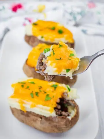 A close-up of Shepherd’s Pie Baked Potatoes topped with ground beef, melted cheddar cheese, and chopped parsley on a white plate. A fork holds a piece with all toppings.