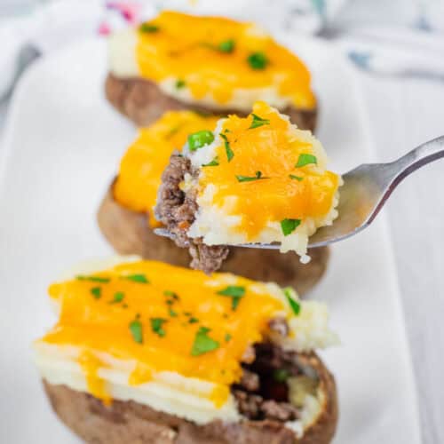 A close-up of Shepherd&rsquo;s Pie Baked Potatoes topped with ground beef, melted cheddar cheese, and chopped parsley on a white plate. A fork holds a piece with all toppings.