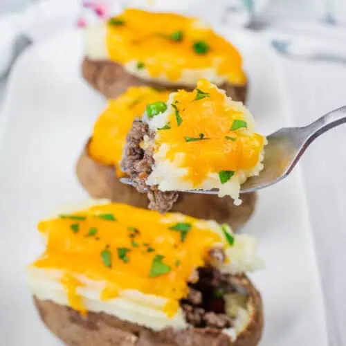 A close-up of Shepherd’s Pie Baked Potatoes topped with ground beef, melted cheddar cheese, and chopped parsley on a white plate. A fork holds a piece with all toppings.
