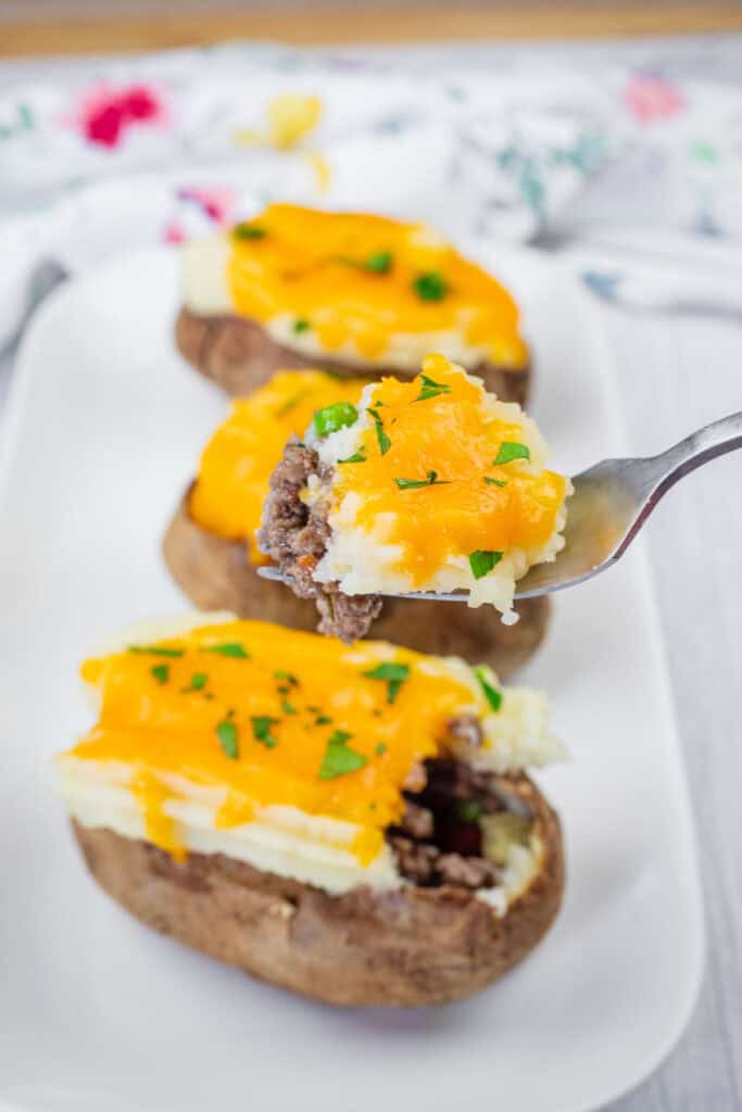 A close-up of Shepherd’s Pie Baked Potatoes topped with ground beef, melted cheddar cheese, and chopped parsley on a white plate. A fork holds a piece with all toppings.
