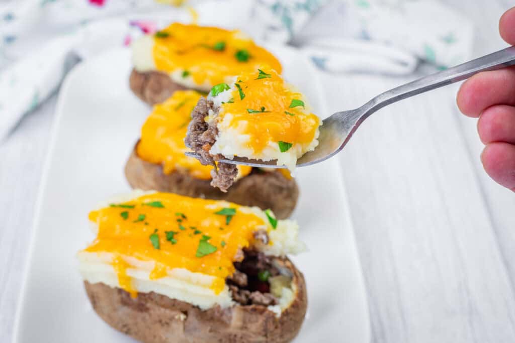 A fork holds a bite of Shepherd&rsquo;s Pie Stuffed Baked Potatoes on a white plate with additional loaded potatoes in the background.