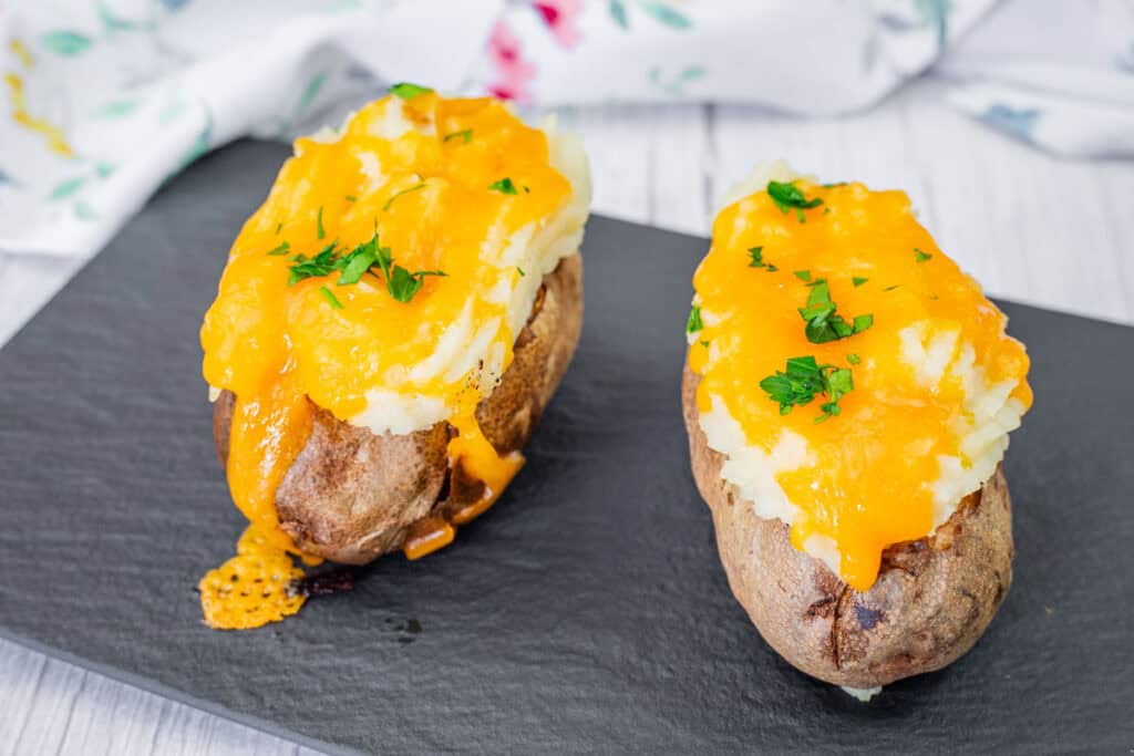 Two Shepherd&rsquo;s Pie Baked Potatoes topped with melted cheddar cheese and chopped parsley on a black tray.