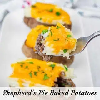 A close-up of Shepherd’s Pie Baked Potatoes topped with ground beef, melted cheddar cheese, and chopped parsley on a white plate. A fork holds a piece with all toppings.
