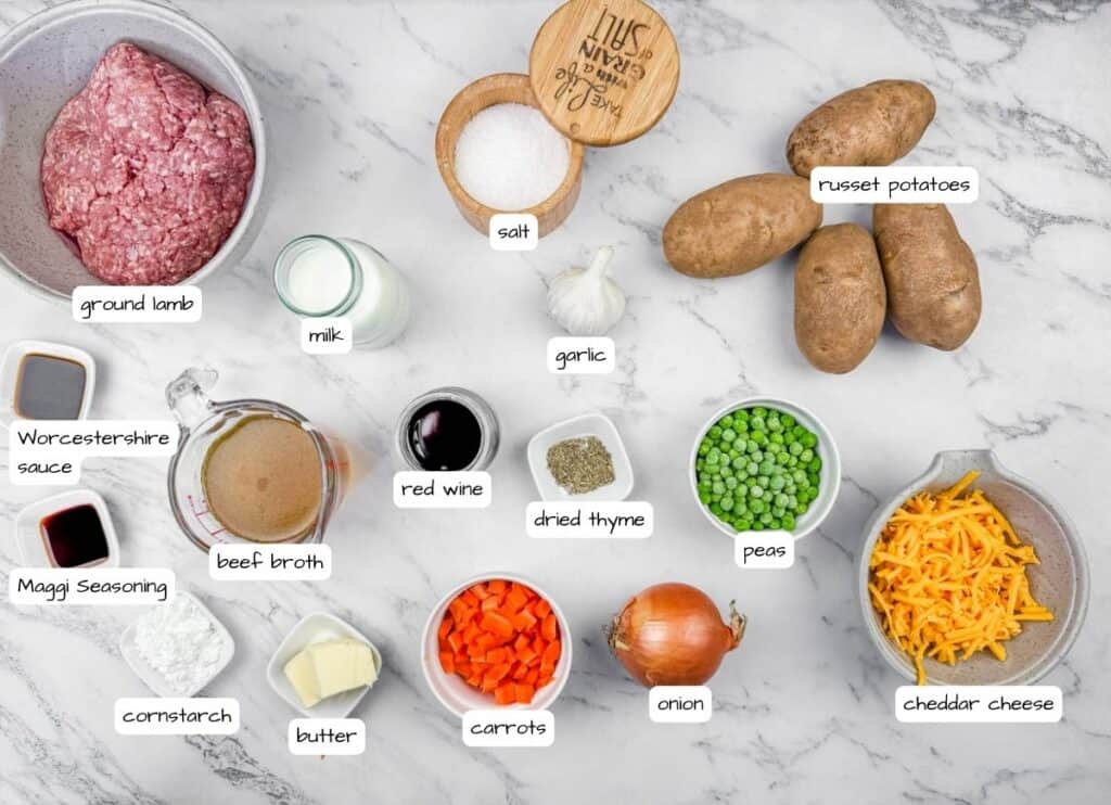Ingredients for Shepherd&rsquo;s Pie Baked Potatoes displayed on a marble surface, including ground lamb, potatoes, peas, carrots, onion, garlic, cheddar cheese, milk, butter, red wine, broth, seasonings, and salt.