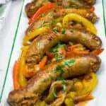 Slow Cooker Sausage & Peppers on a rectangular plate with sliced red, yellow, and green bell peppers, garnished with chopped herbs.