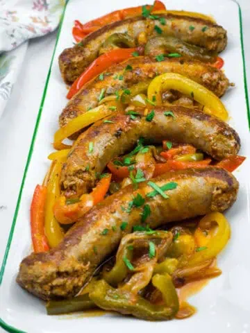 Slow Cooker Sausage & Peppers on a rectangular plate with sliced red, yellow, and green bell peppers, garnished with chopped herbs.