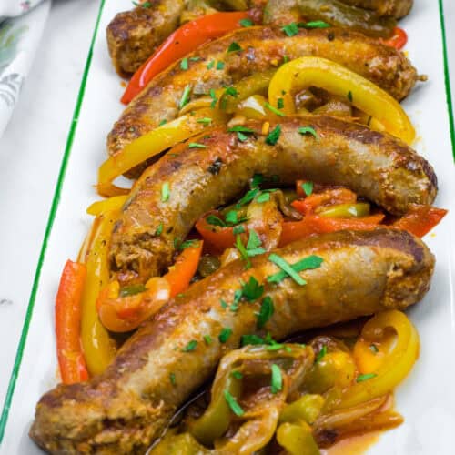 Slow Cooker Sausage & Peppers on a rectangular plate with sliced red, yellow, and green bell peppers, garnished with chopped herbs.
