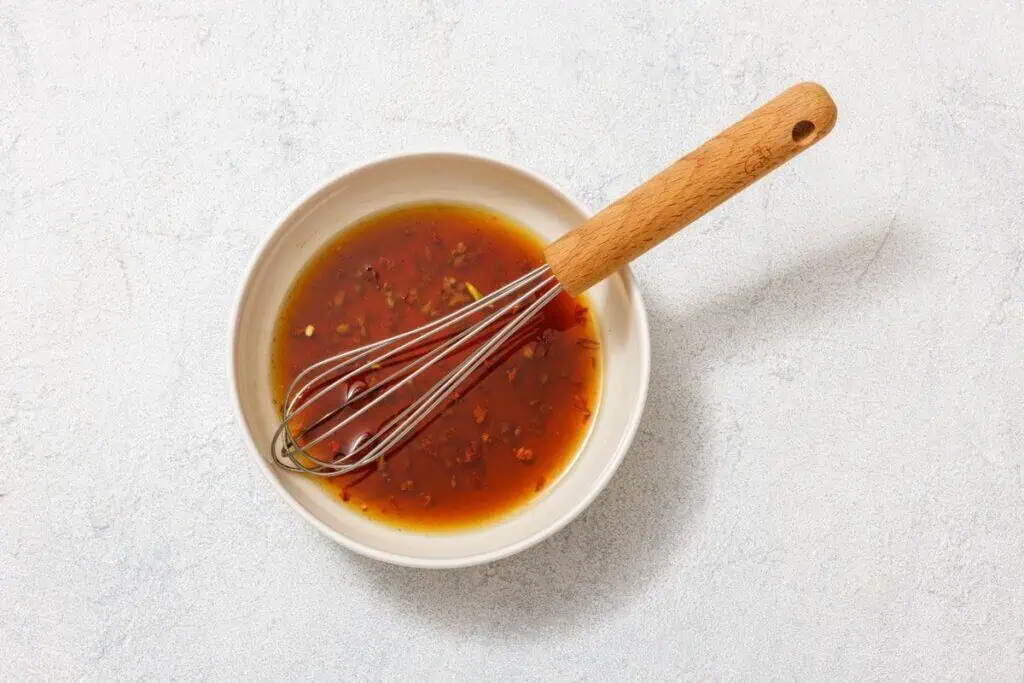 Bowl of brown sauce with chopped herbs, garlic, and spices, accompanied by a whisk, on a light textured surface.