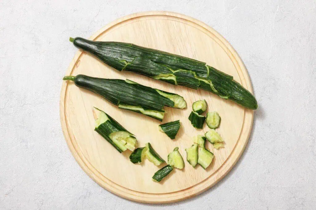 Two partially peeled cucumbers on a round wooden board, with additional pieces cut and scattered around.